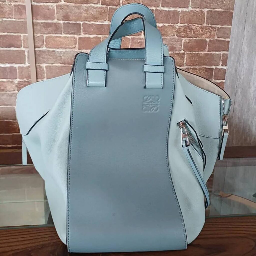 Light blue leather backpack with top handles and a front panel logo, resting on a wooden surface against a brick wall.