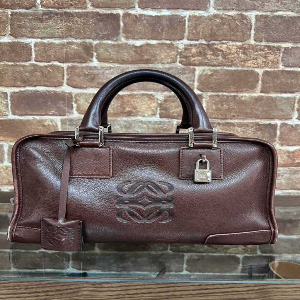 Brown leather handbag with rounded top handles, embossed logo on front, silver hardware, and a matching luggage tag against a brick wall.