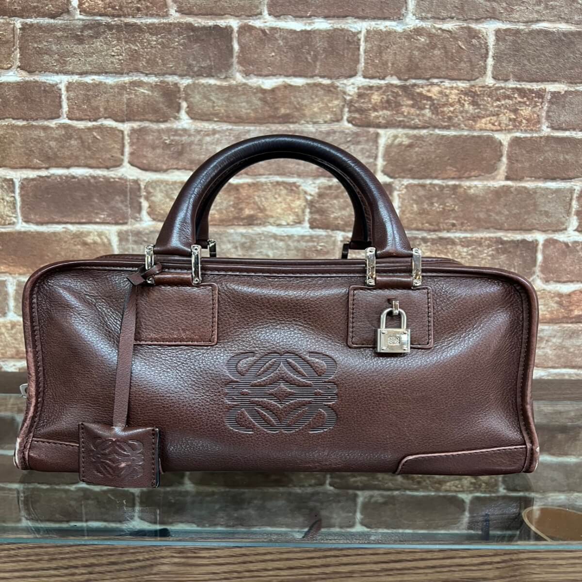 Brown leather handbag with rounded top handles, embossed logo on front, silver hardware, and a matching luggage tag against a brick wall.