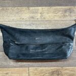 Black leather crossbody bag with a front zip pocket and small brand tag, lying on a wooden floor underneath a zipper at the top edge.