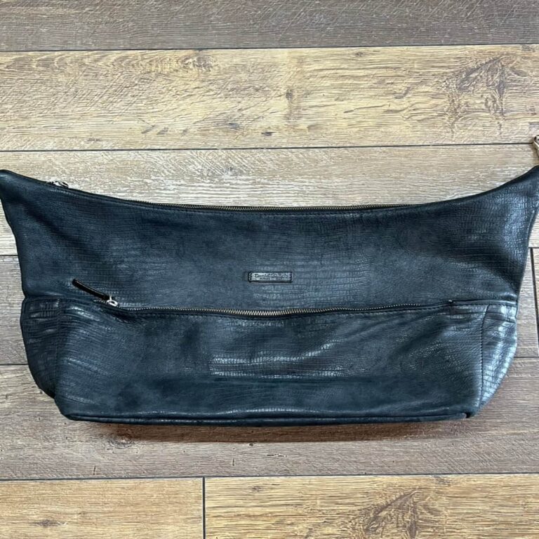 Black leather crossbody bag with a front zip pocket and small brand tag, lying on a wooden floor underneath a zipper at the top edge.