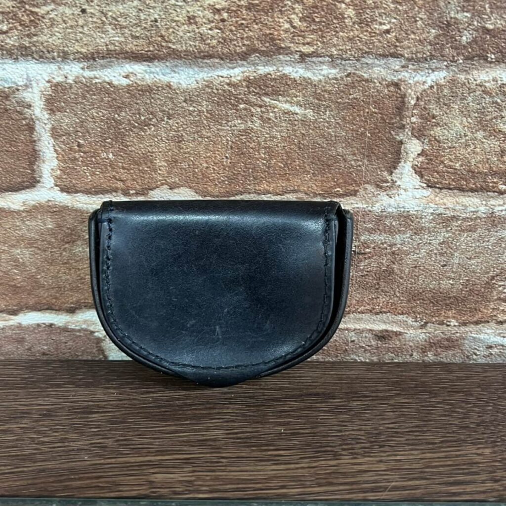 Small black leather coin purse resting on a wooden surface against a brick wall; half-moon shape with visible stitching.