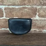 Small black leather coin purse resting on a wooden surface against a brick wall; half-moon shape with visible stitching.