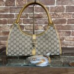 Designer beige handbag with the GG monogram and tan leather trim displayed against a brick wall, on a stand with a small accessory in plastic nearby.
