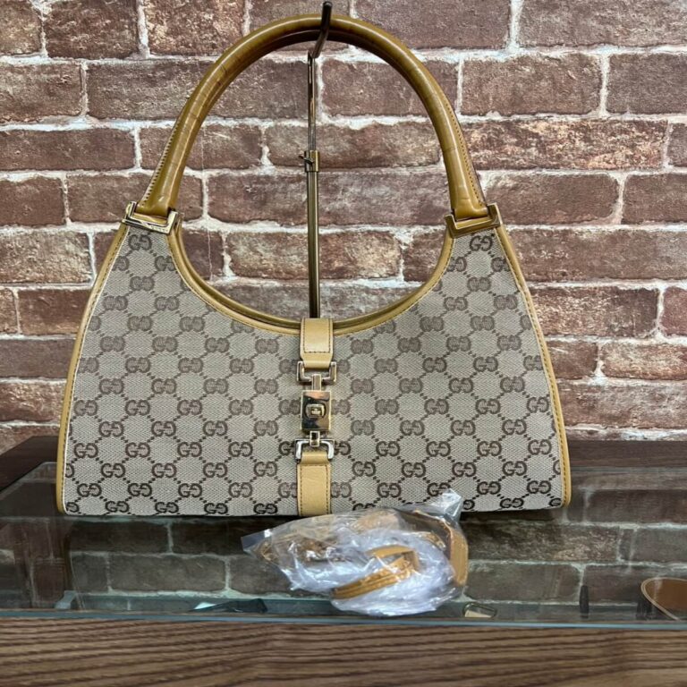 Designer beige handbag with the GG monogram and tan leather trim displayed against a brick wall, on a stand with a small accessory in plastic nearby.
