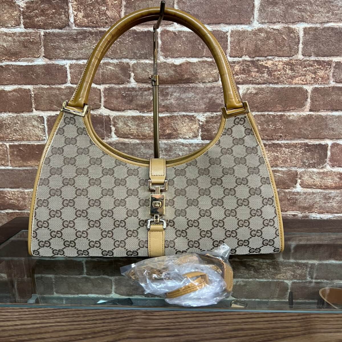 Designer beige handbag with the GG monogram and tan leather trim displayed against a brick wall, on a stand with a small accessory in plastic nearby.
