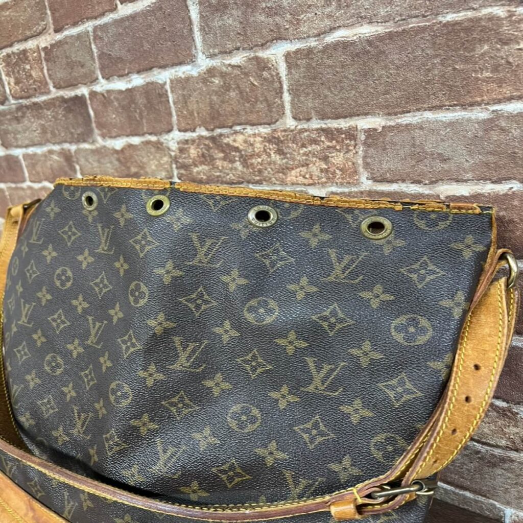 Louis Vuitton monogram handbag with brass grommets and tan leather strap, resting against a brick wall, showing wear on the trim.