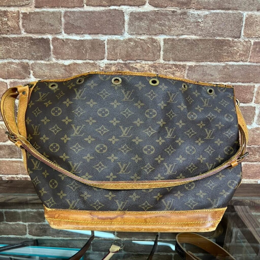 Louis Vuitton monogram canvas shoulder bag with brown leather trim and metal grommets against a brick wall backdrop.