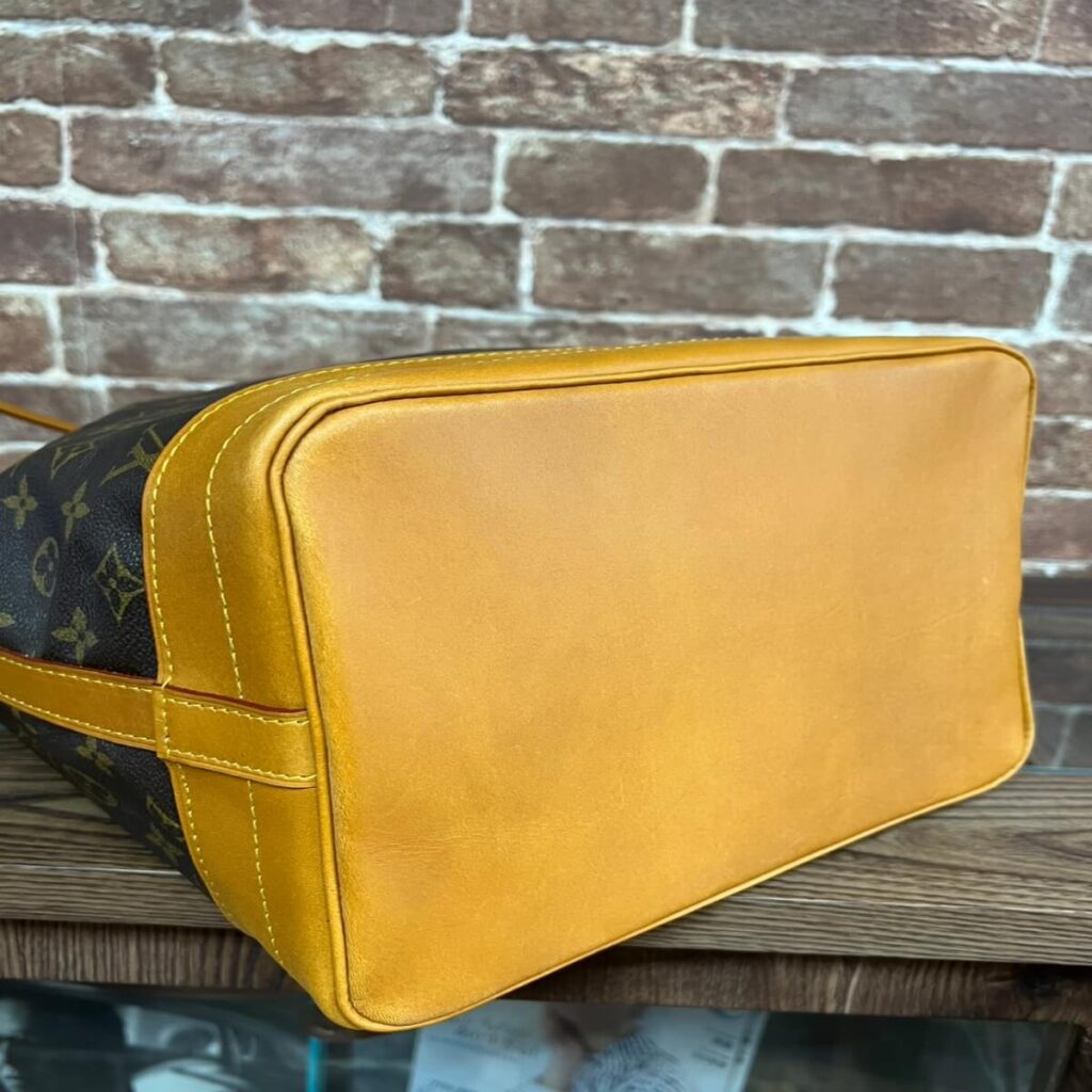 Bottom view of a designer handbag with mustard-yellow leather base, visible stitching, and a monogrammed dark canvas side.