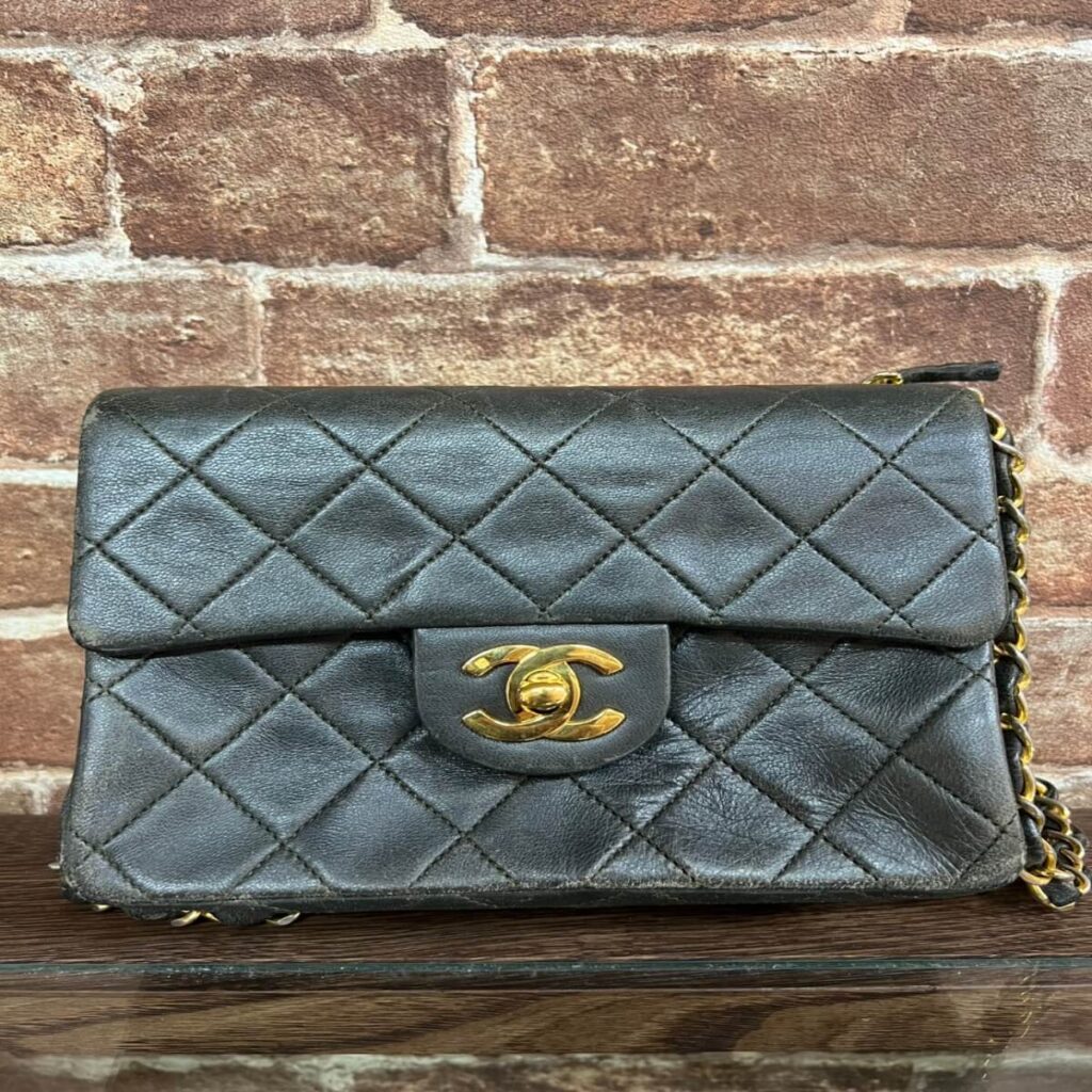 Black quilted Chanel handbag with gold-tone interlocking CC turn-lock, chain strap visible against brick wall surface, classic flap design.