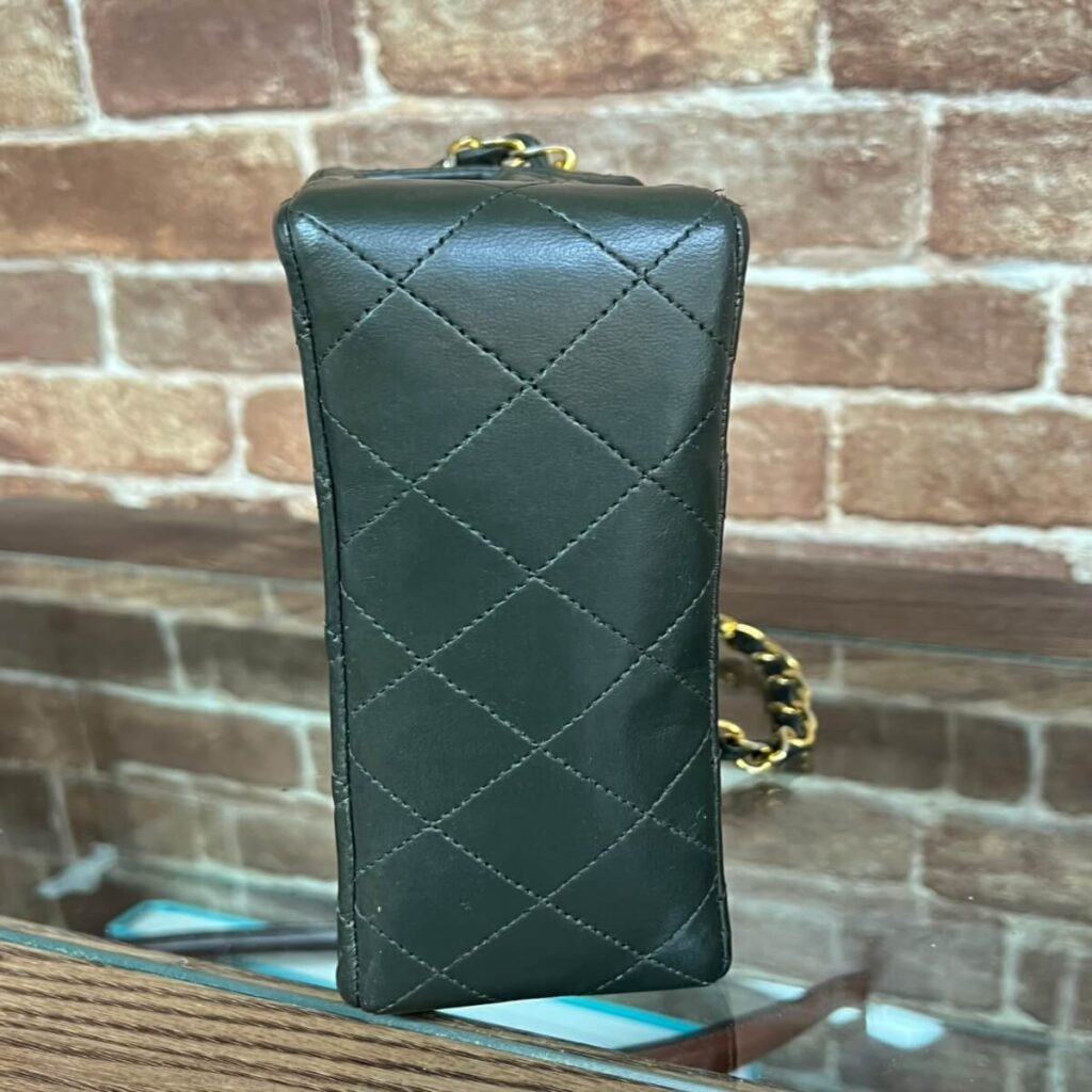 Black quilted leather handbag with a gold chain strap, standing on a glass surface against a brick wall backdrop