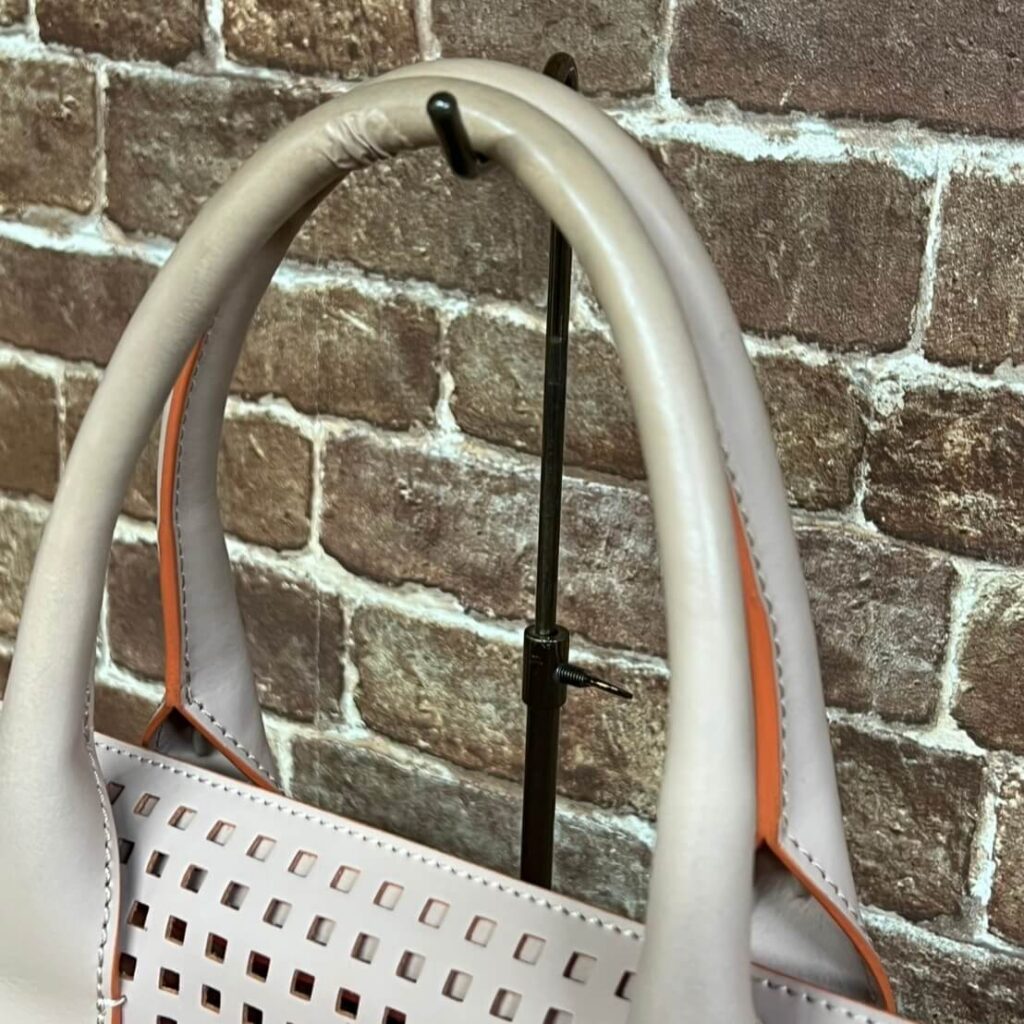 Light gray handbag with orange piping on a display stand against a brick wall, showing square perforations on the bag body