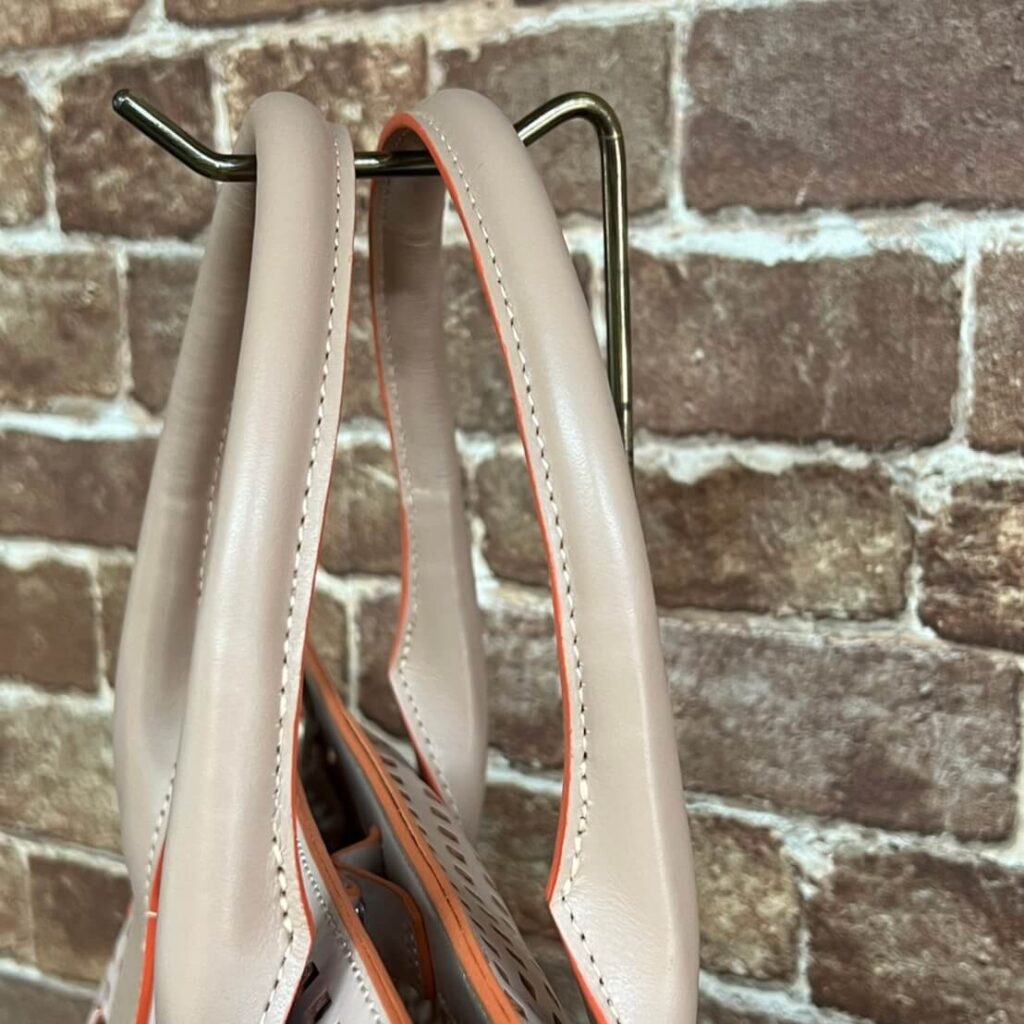 Beige leather handbag with orange piping hanging on a metal rack against a brick wall.