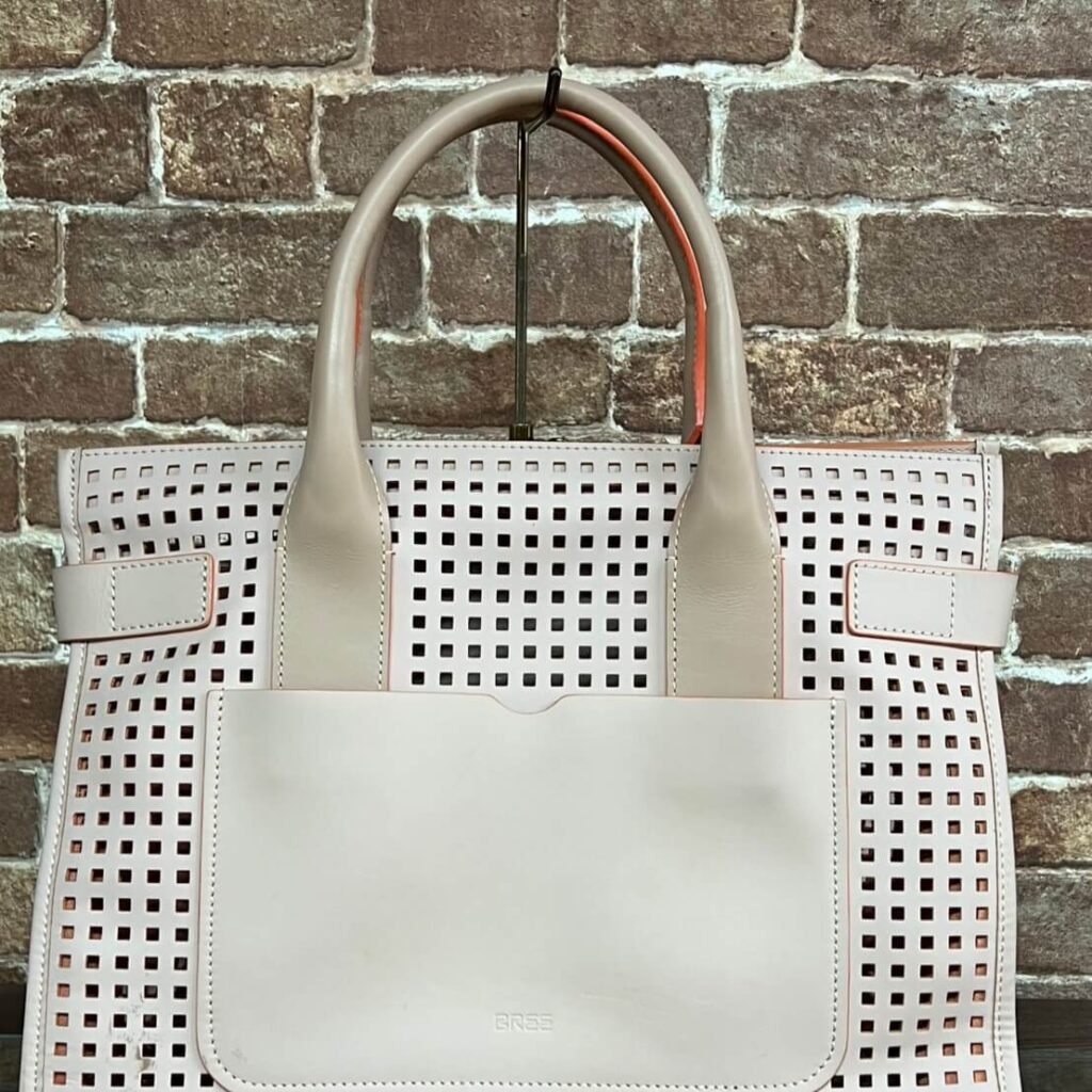 Beige perforated tote bag with square holes and a front pocket, hanging on a stand against a brick wall.