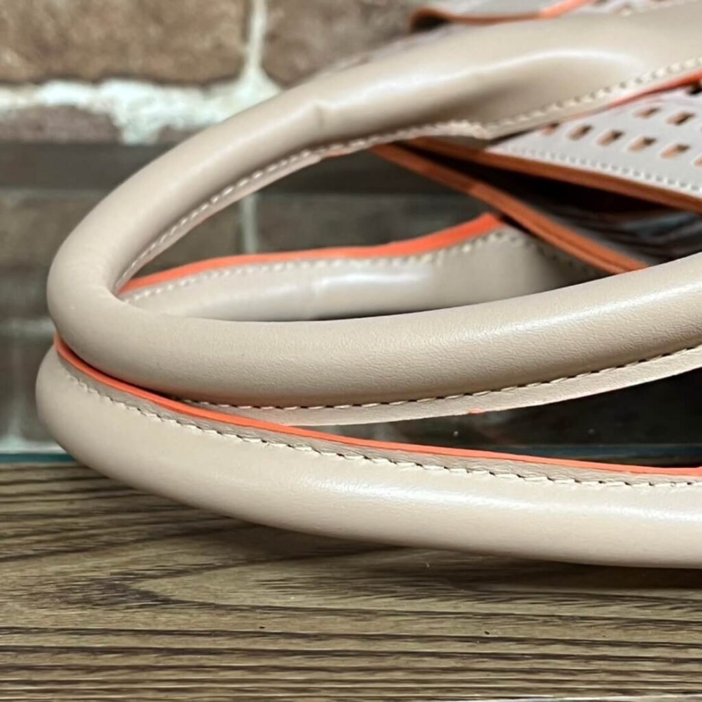 Close-up of a beige leather bag strap with orange piping resting on a wooden surface.
