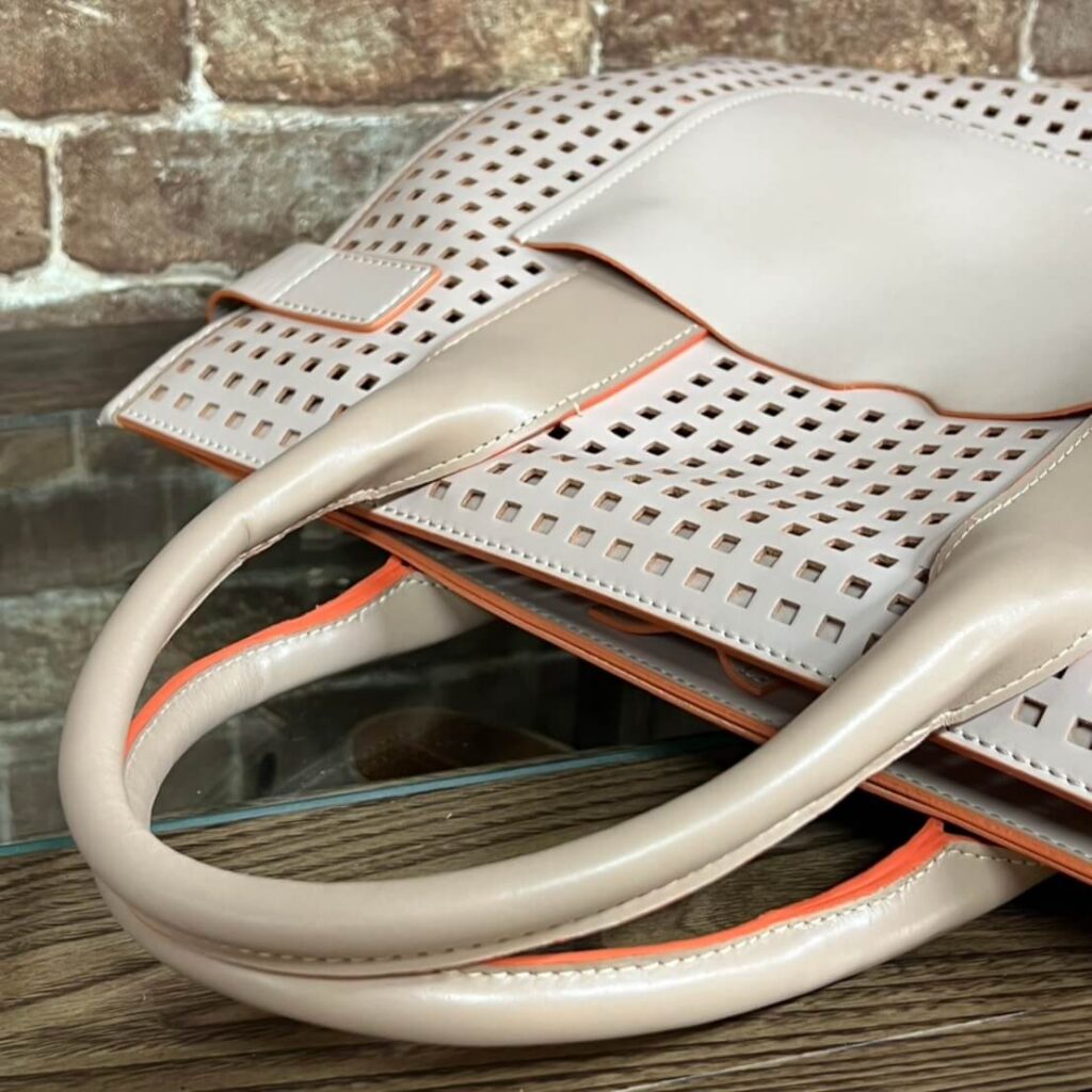 Beige perforated handbag with orange trim and curved handles resting on a wooden table by a brick wall.