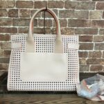 White perforated tote bag with a large front pocket, pink edging, and rounded handles against a brick wall.