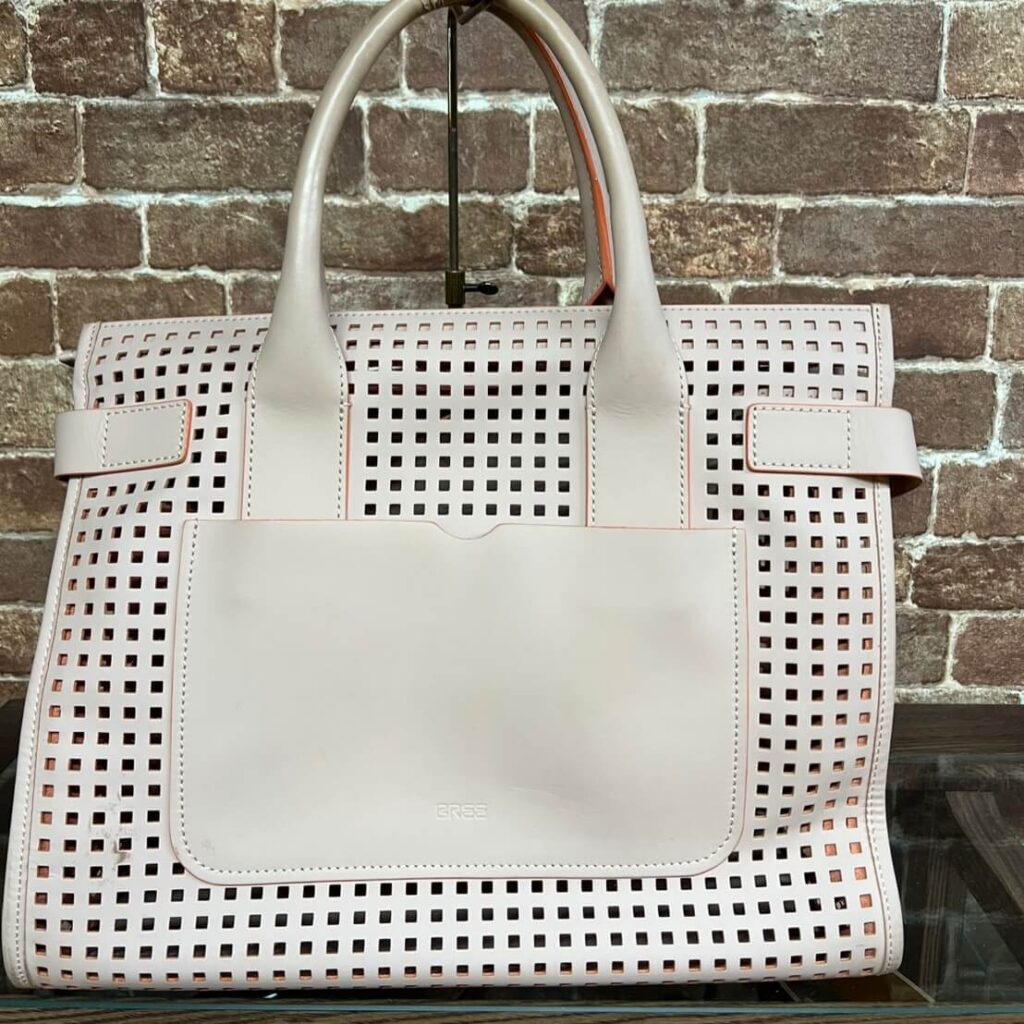 White perforated leather tote bag with square cutouts and a front pocket, two handled, against a brick wall.